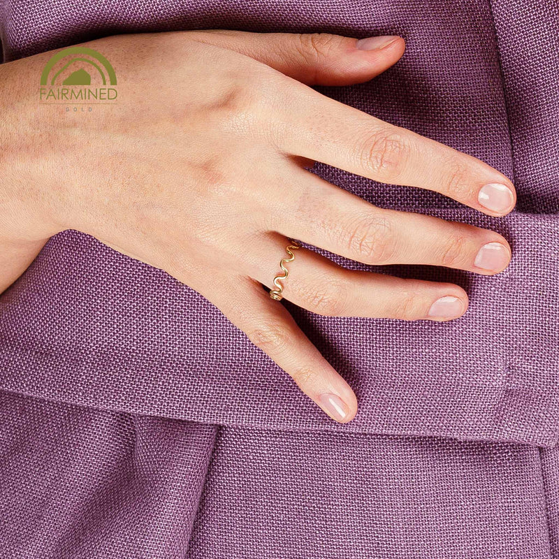 Hand wearing a  wavy gold ring on a purple fabric background with Fairmined logo.