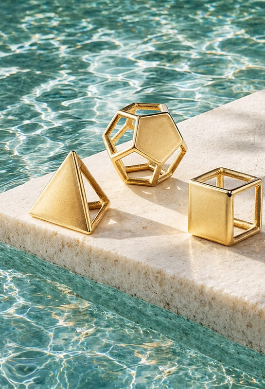Three gold geometric jewels on a stone ledge by a pool.